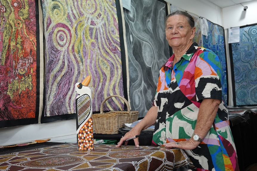 A photo of a woman wearing printed shirt standing next to Indigenous artpieces