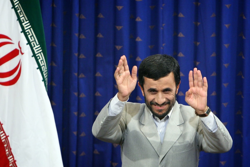 A middle-aged Iranian man with a beard in a grey suit smiles and waves in front of an Iranian flag.