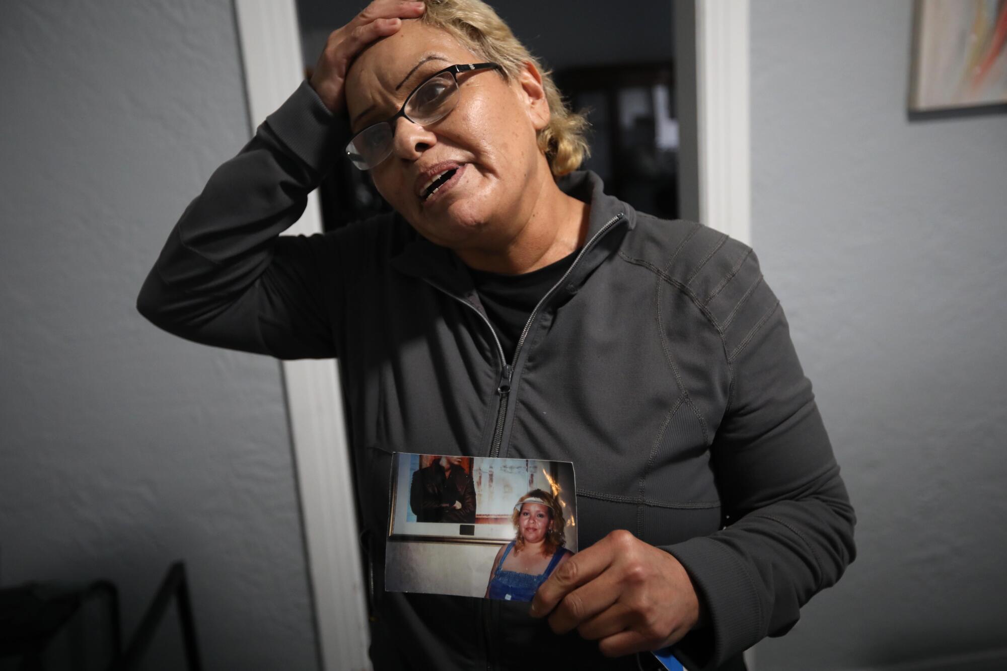 A woman puts her right hand on her head while holding a photo of her younger self.