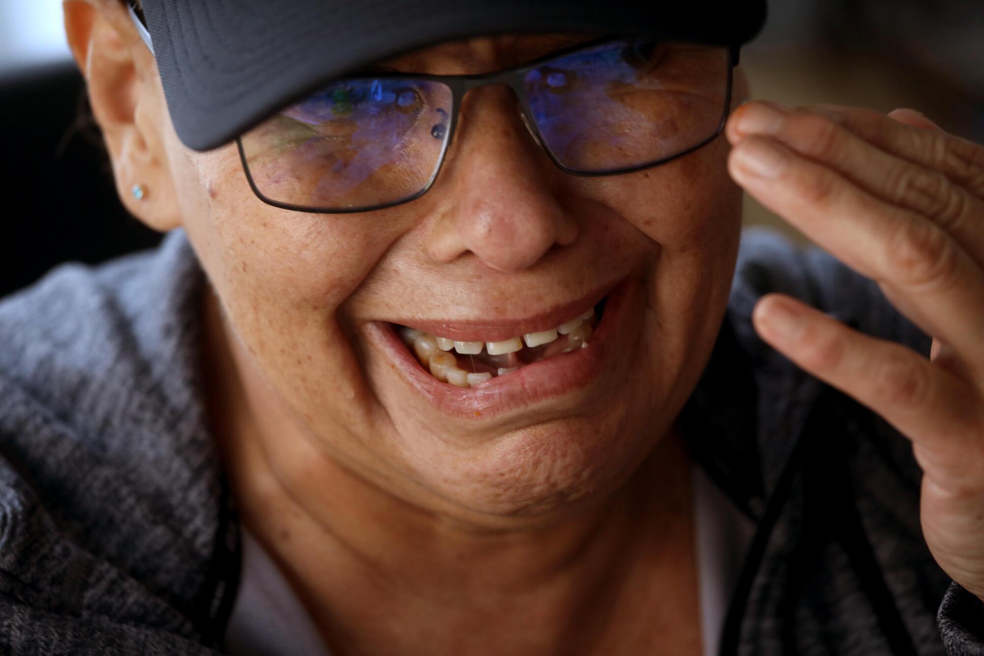 A woman wearing glasses and a cap cries while recalling all the hardships and loss she has suffered over the years.