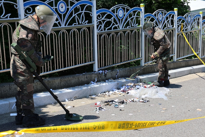 Two soldiers in protective equipment scan trash sitting in a street next to a fence