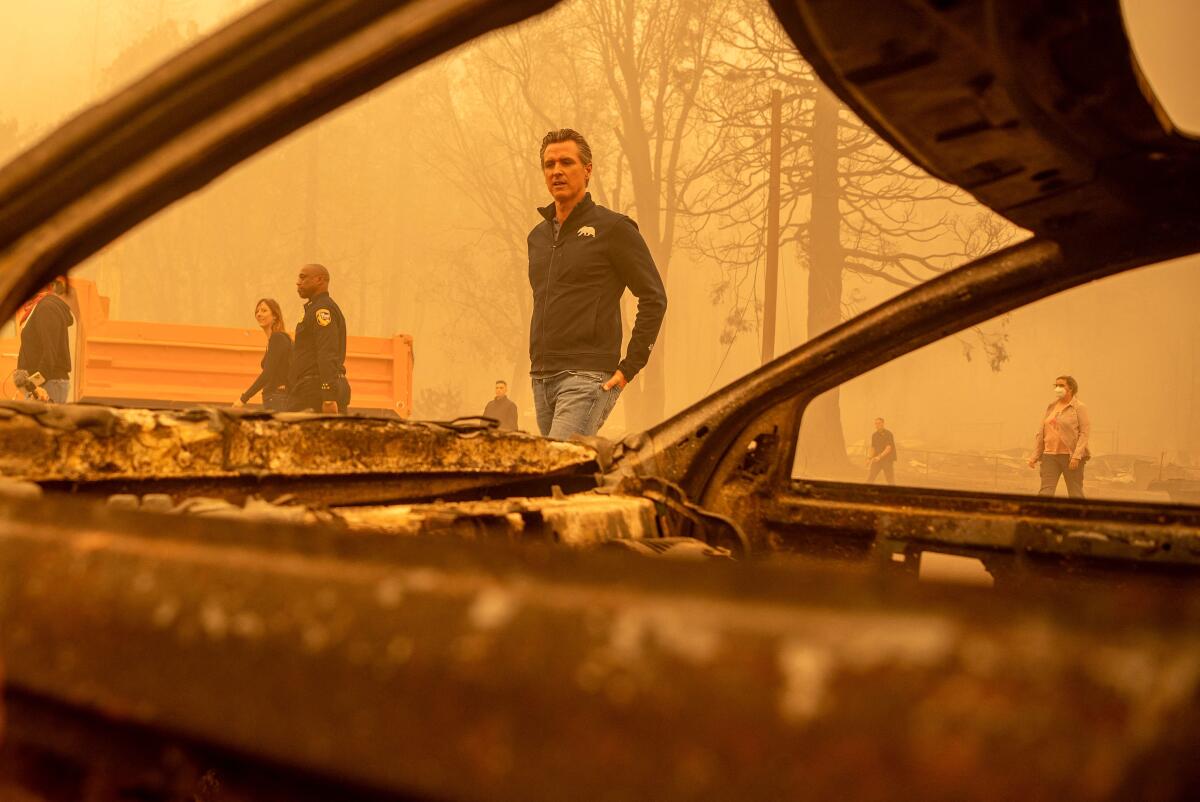 Gov. Gavin Newsom is framed by a burned vehicle. The sky is orange with wildfire smoke.