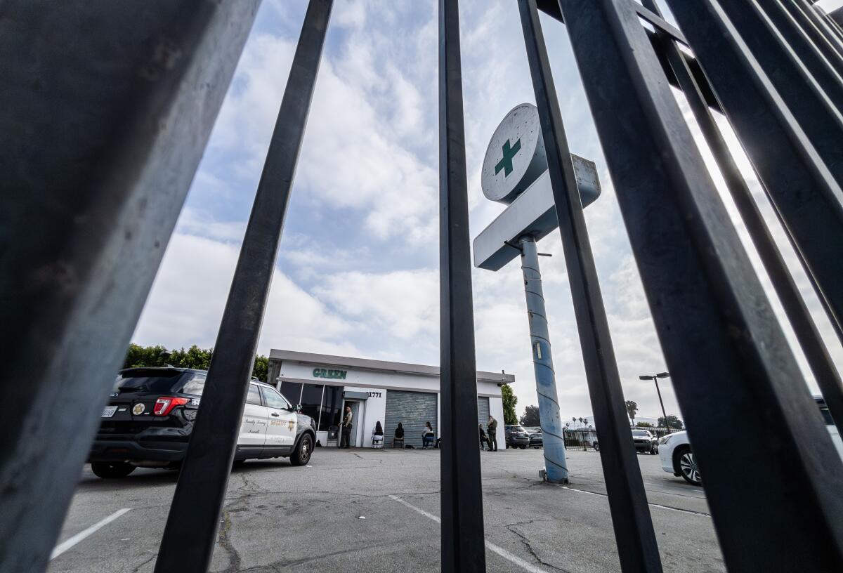 A photo taken through metal bars shows a Sheriff's Department SUV parked near a cannabis dispensary and its green cross sign