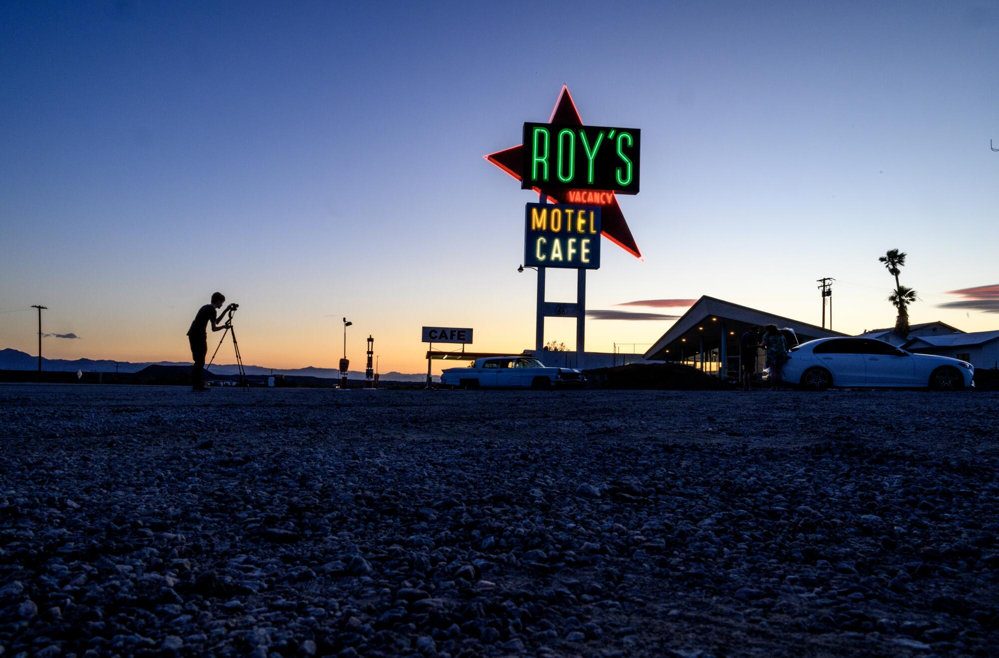 The Roy's Motel and Cafe neon sign glows against the dusk, with a person and camera tripod silhouetted.