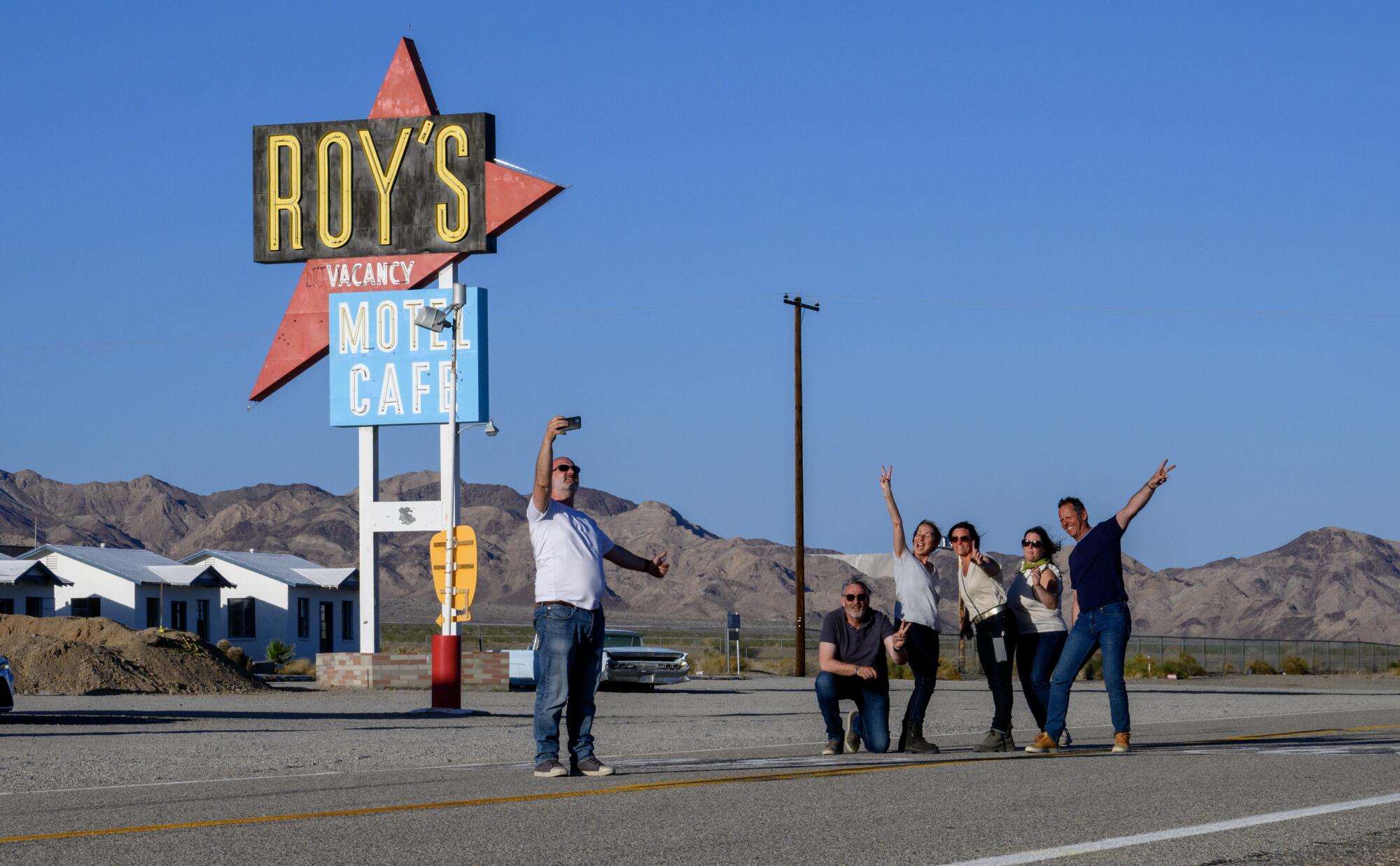 A group of people stand in the road on Route 66, posing for a selfie with the Roy's sign.