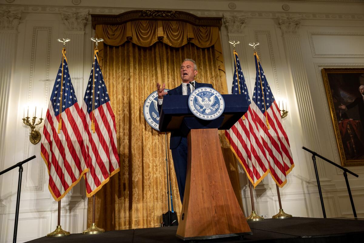 Kennedy speaks from a stage with flags behind him 