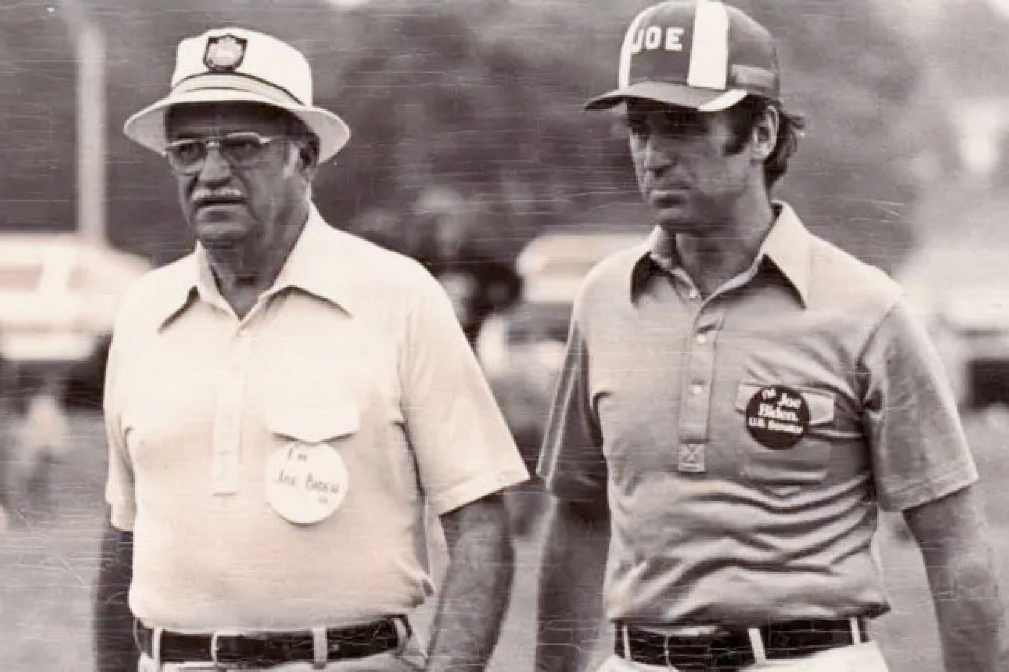 A sepia-colored photo of a man wearing glasses and a hat next to a younger man wearing a cap.