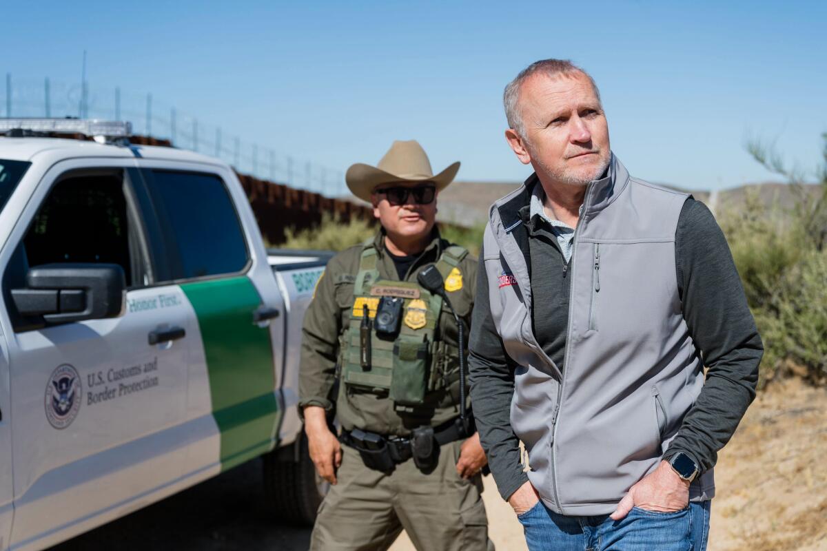 Matt Gunderson and a man in a green uniform and tan cowboy hat looking around by a white government pickup near a border wall
