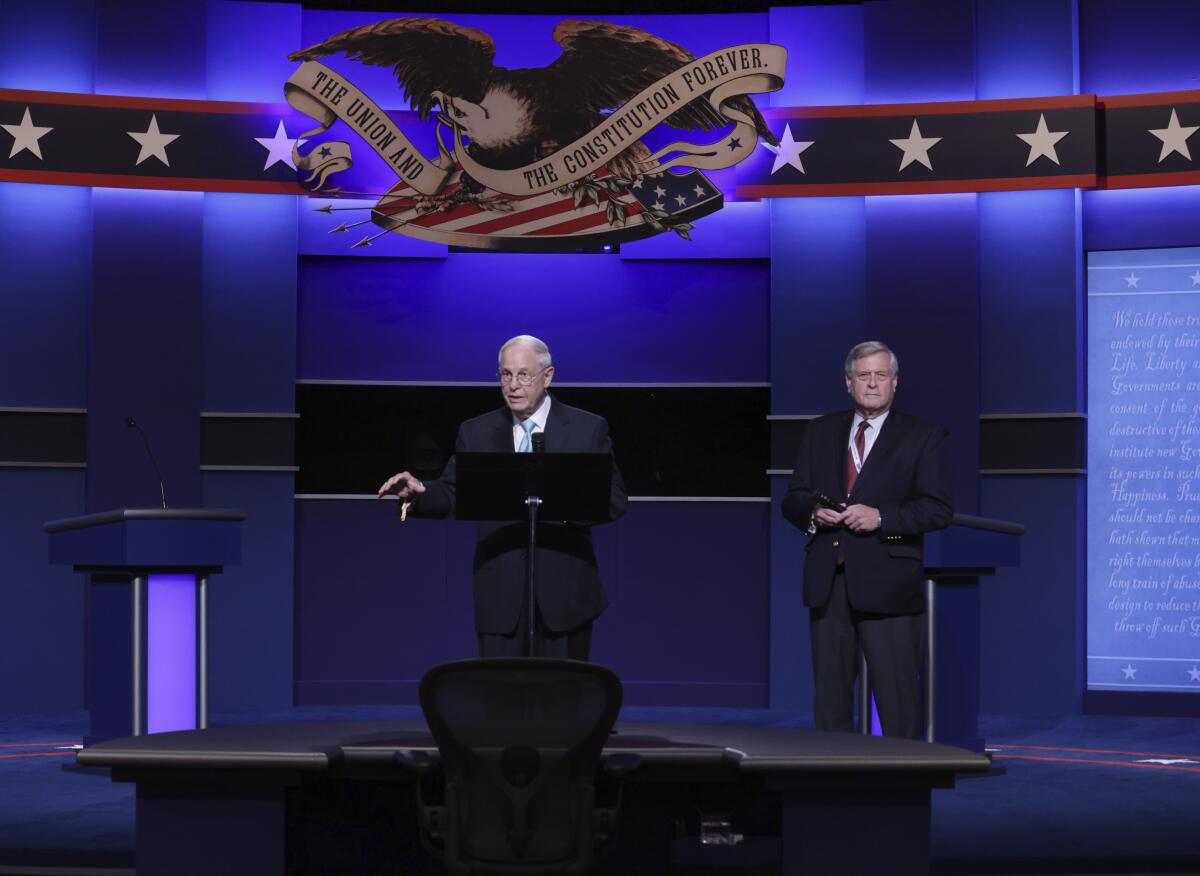 Frank Fahrenkopf and Kenneth Wollack stand on a presidential debate stage with patriotic decor and a bright blue background