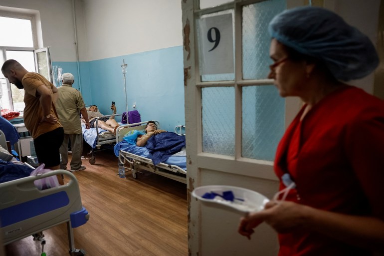 A nurse entering a hospital ward where four injured people are lying on their beds. She is carrying a dish containing medical equipment. Two of the injured have visitors.