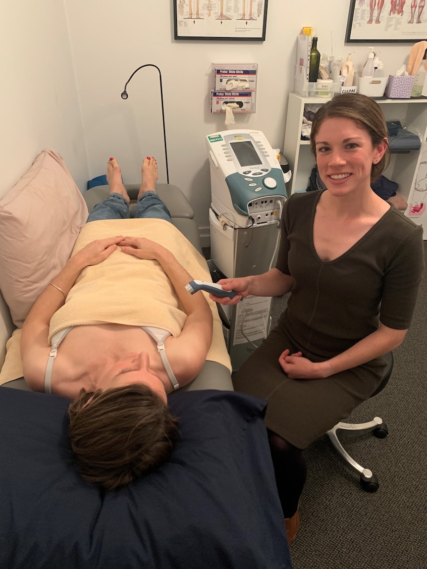 Emma Heron sits beside a patient in her clinic and holds an ultrasound wand, smiling at the camera.