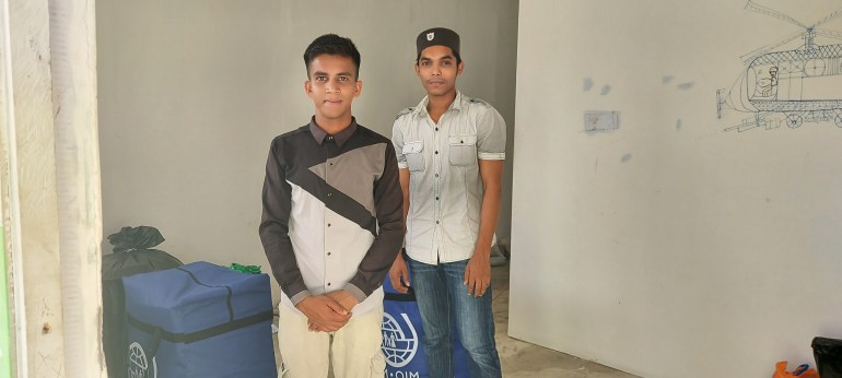 Gura Amin and M Ullah as they prepare to leave Aceh. There are large blue carriers with IOM written on them and a logo. On the wall beside them is a drawing of a helicopter