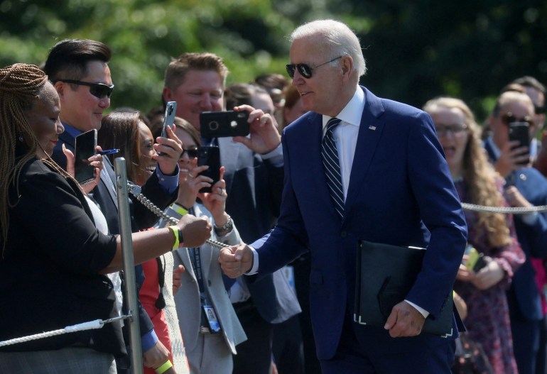 Joe Biden walks along a line of supporters, who stand behind a barricade outside, some pointing camera phones at the president.