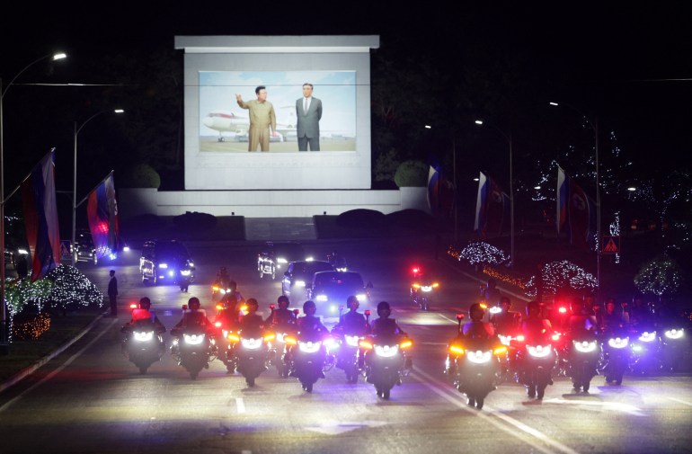The motorcade taking Putin and Kim into the city. Outriders in formation are in front. It is dark so the vehicles all have their lights on. There is a large billboard behind depicting Kim Jong-il and Kim Il-sung