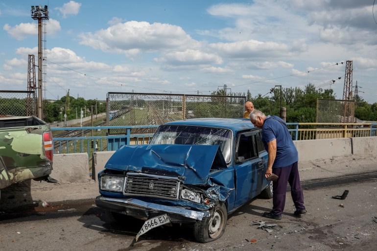 A man weeps by the front door of a blue car where his son was killed in a Russian attack. The front of the car is mangled with the number plate hanging off. The windscreen is broken. Another damaged car is nearby.