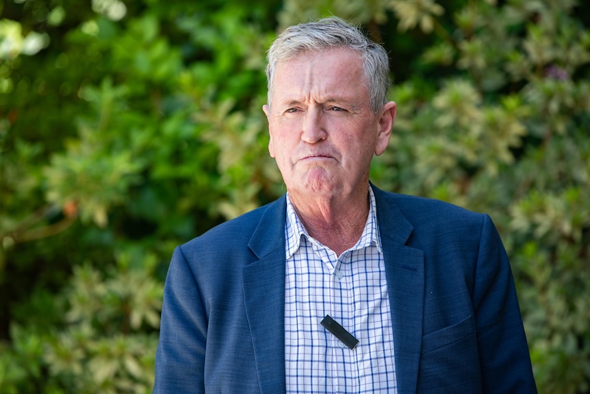 A man in a button up shirt and blazer speaks against a leafy background.