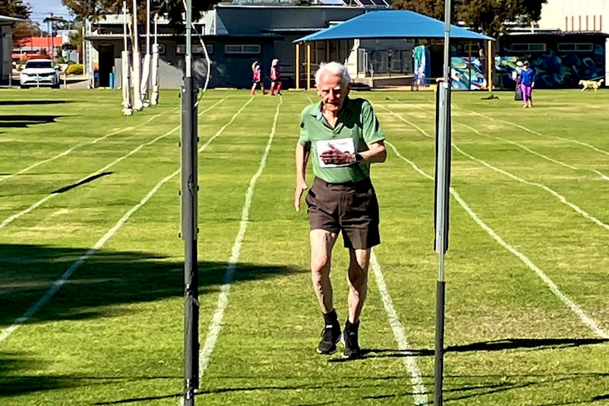 an elderly man in a green top and brown shorts crosses the finishing line at a race on green grass