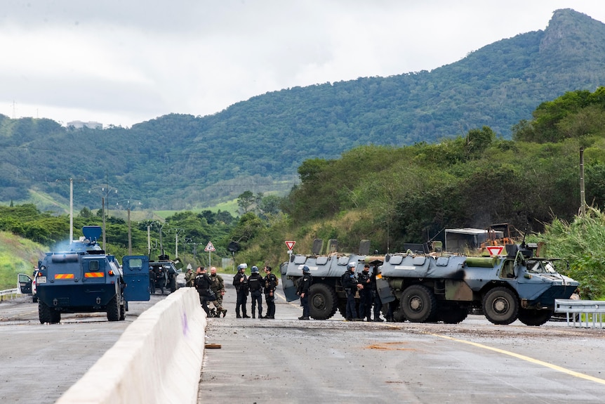 Officers stand next to large blue police vehicles on a highway surrounded by green trees and small mountains