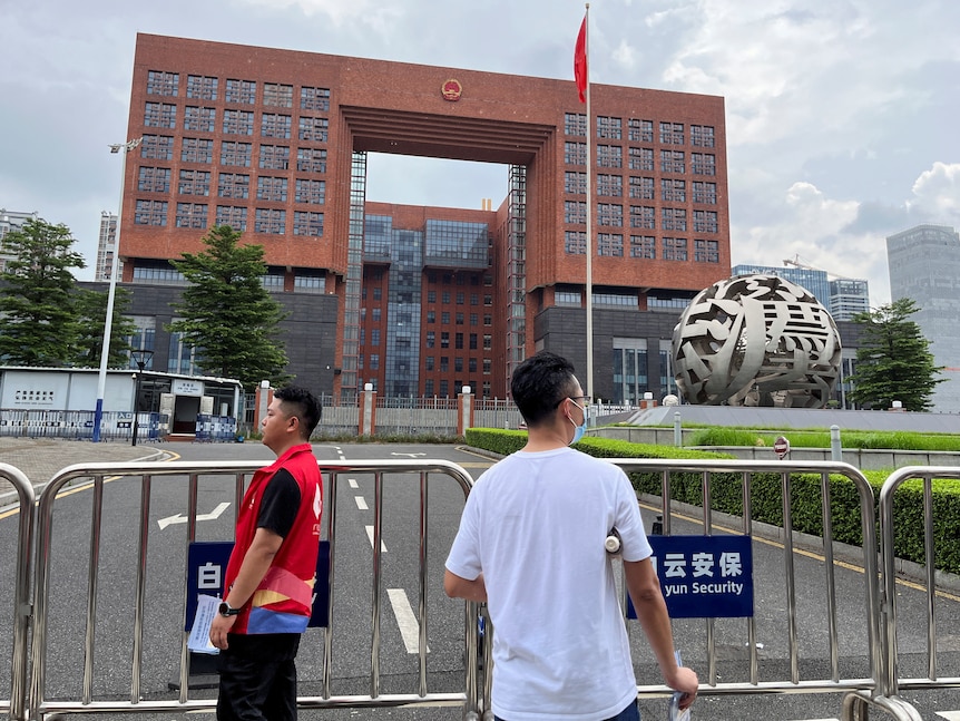 A large red-brick building sits behind a large fence with people watching on in the foreground