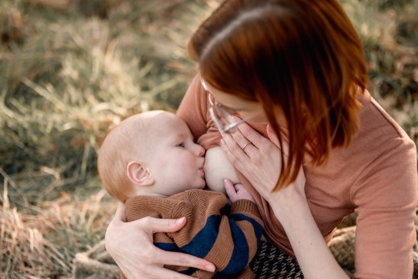 Katya Daniel sits on the grass and lifts her shirt to allow her baby to breastfeed.