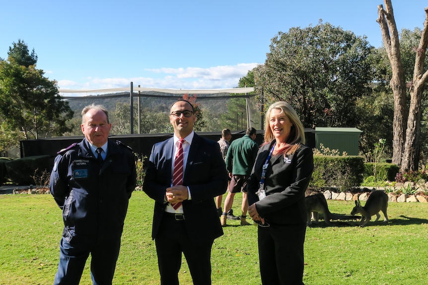 Two men and a woman stand on a lawn in front of kangaroos. 