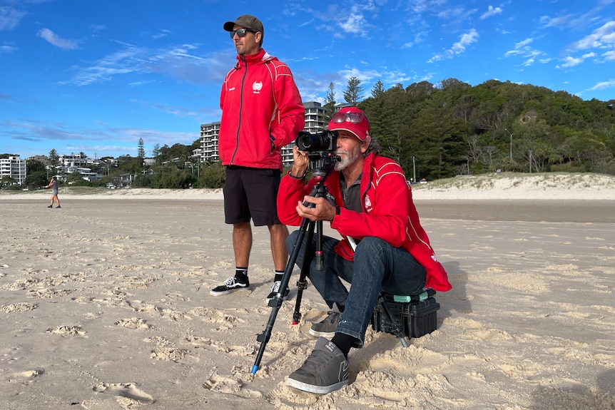 two school surf coaches on the beach one looking down the lense of a camera towards the waves