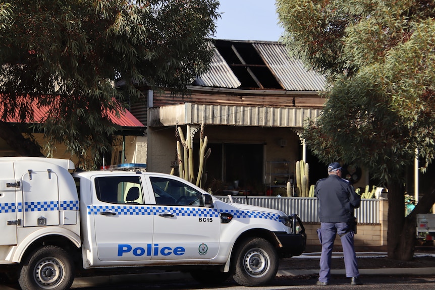 A man in a police uniform standing with his back to the camera next to a police car in front of a house damaged by fire