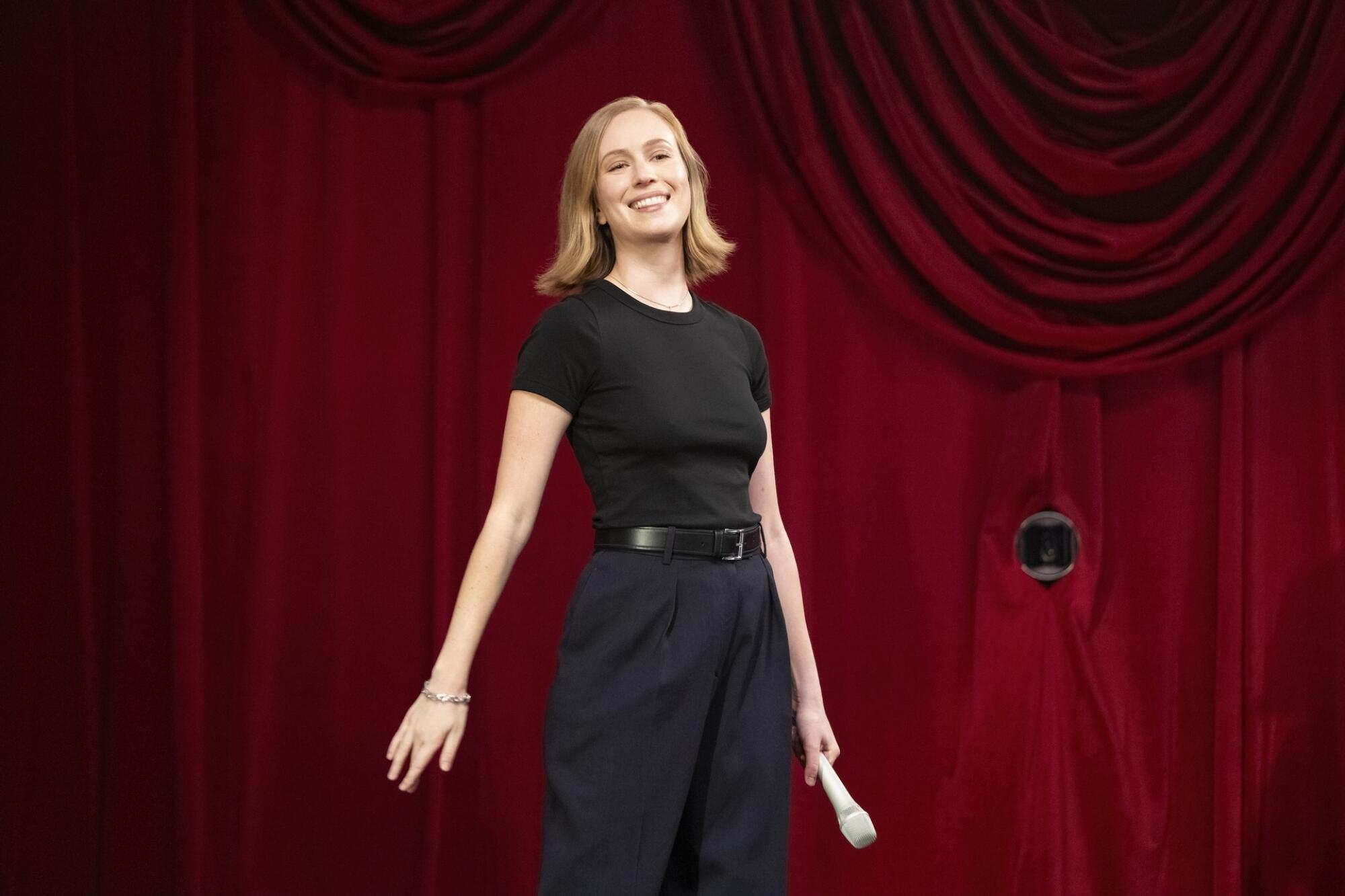 Hannah Einbinder in a black T-shirt and slacks holding a microphone and standing in front of burgundy stage curtains.