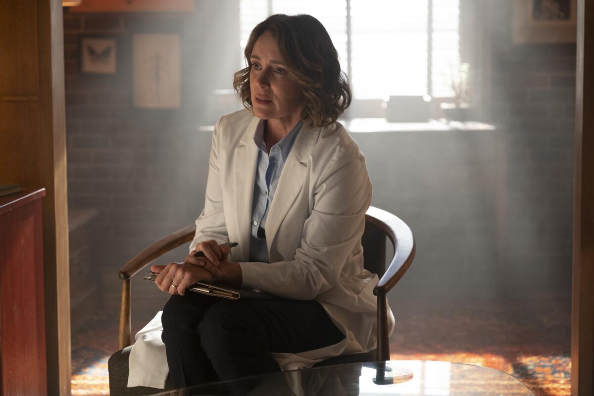 A woman in a white lab coat sits in a chair with her legs and hands crossed.