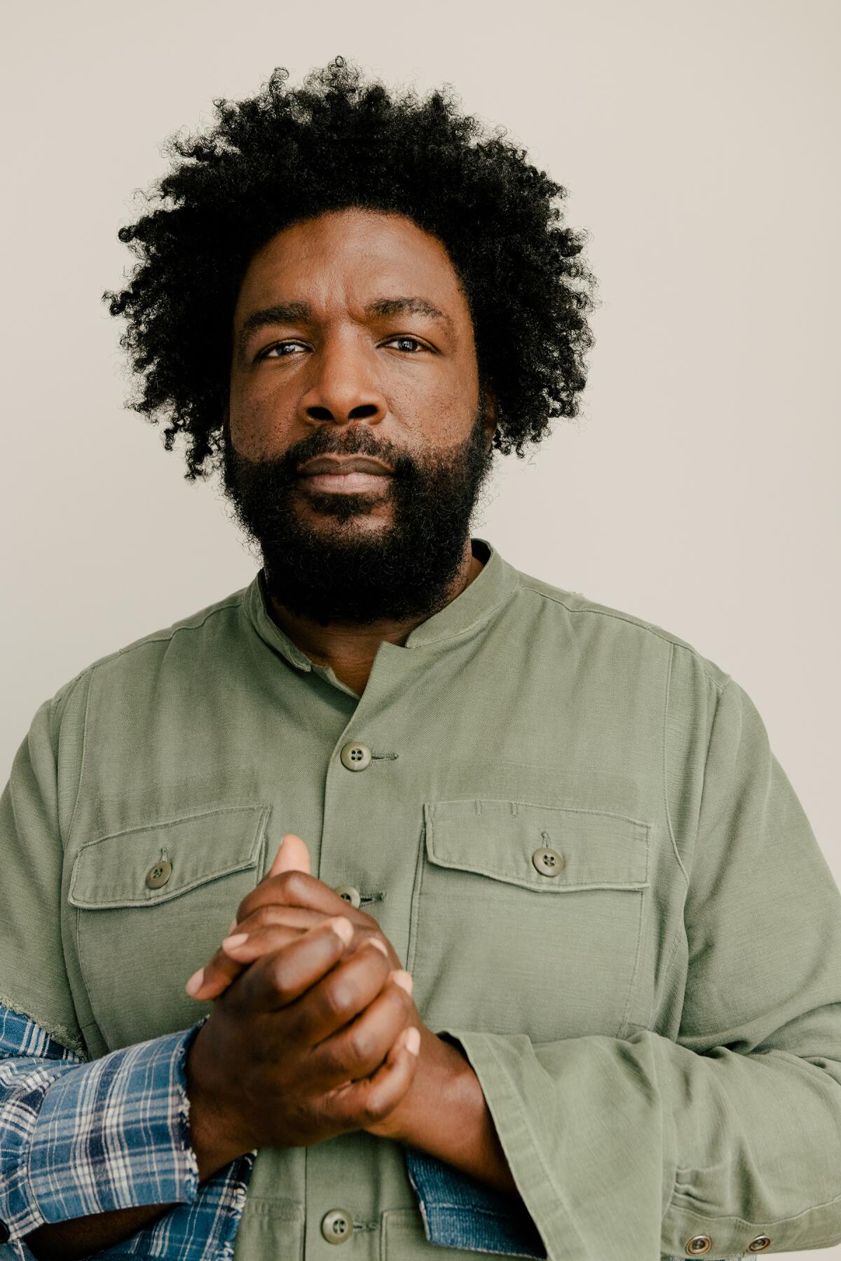 A man with dark hair and beard, in a light green jacket, hands clasped