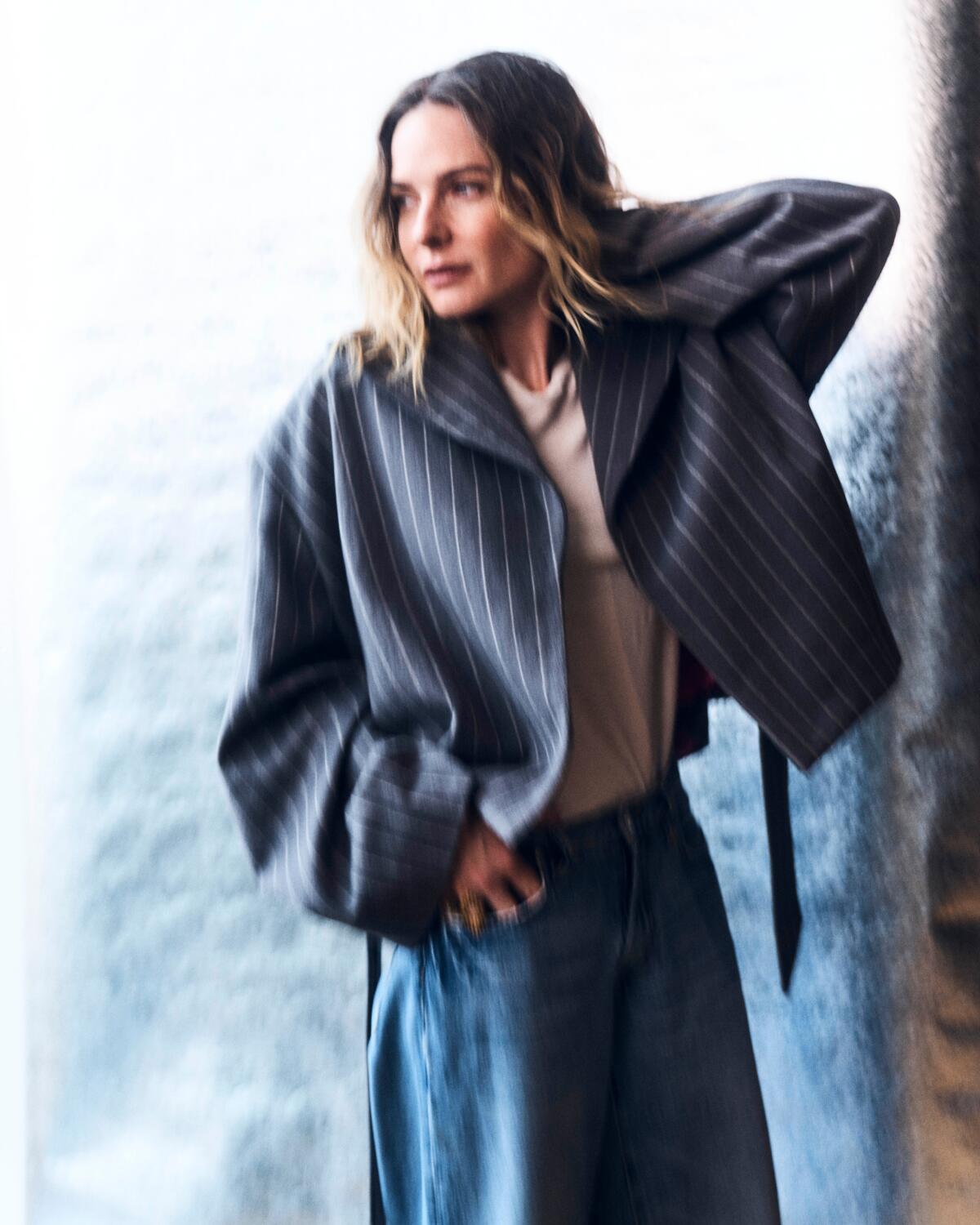 Rebecca Ferguson leans against a wall looking to the side for a portrait.