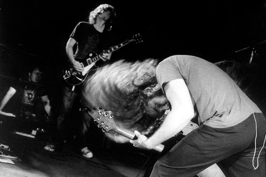 A guitarist head bangs his long hair while a bandmate performs in the background.