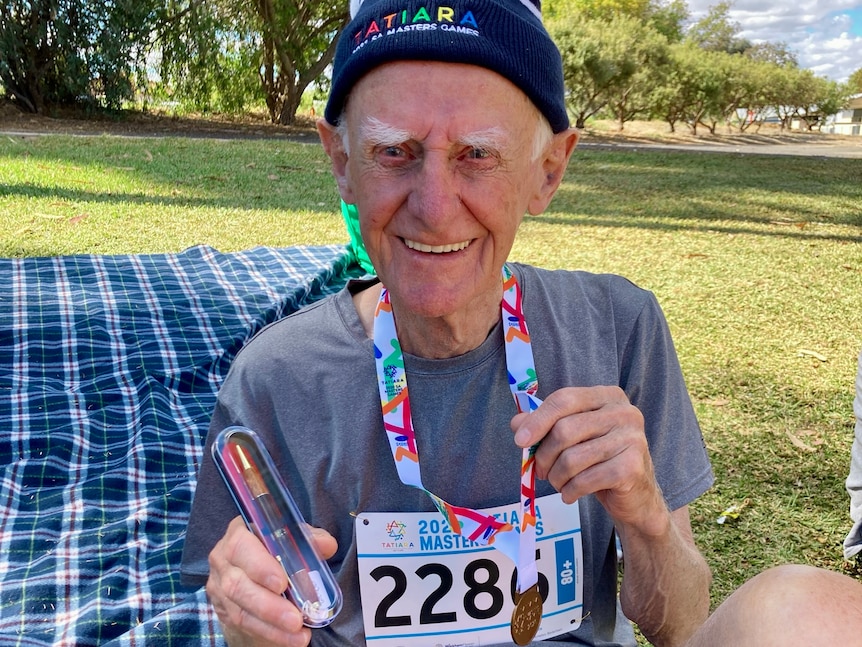 An elderly man smiles, sitting on the grass wearing a blue and white beanie holding a gold medal and a gold pen