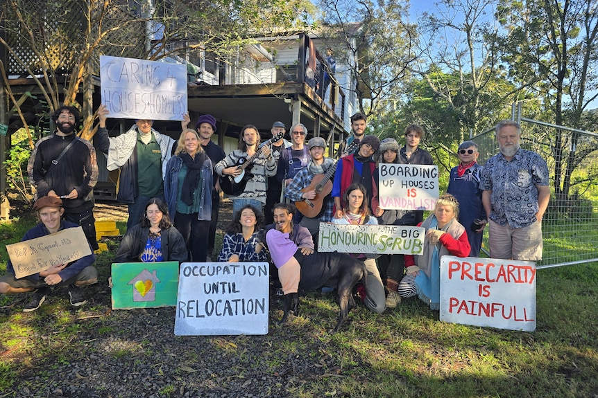 Twenty people with placards stand outside barricaded, flood-affected house.