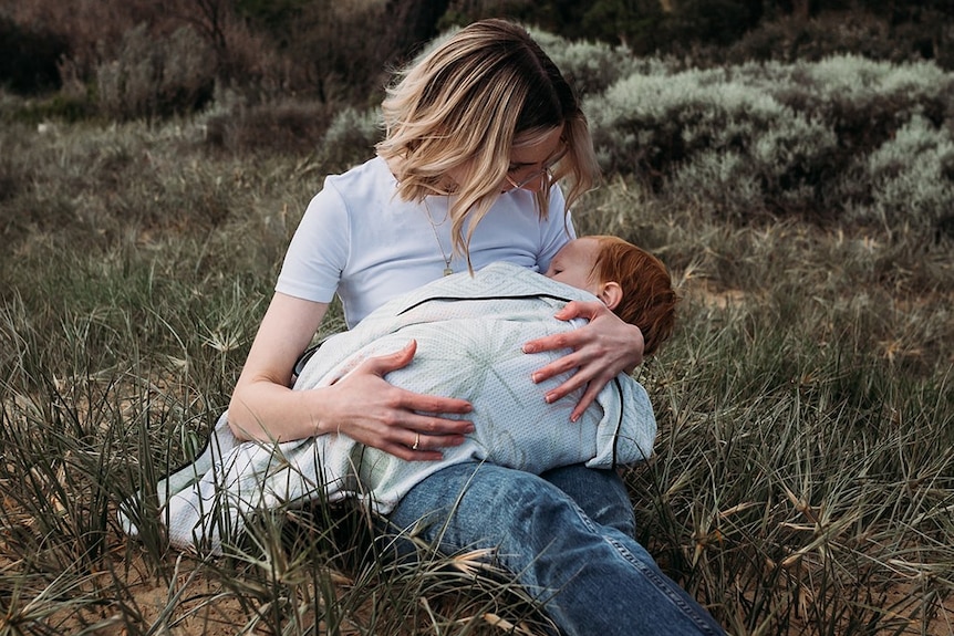 Katya Daniel sits in a grassy field and looks down at her son who is wrapped in a blanket on her lap.