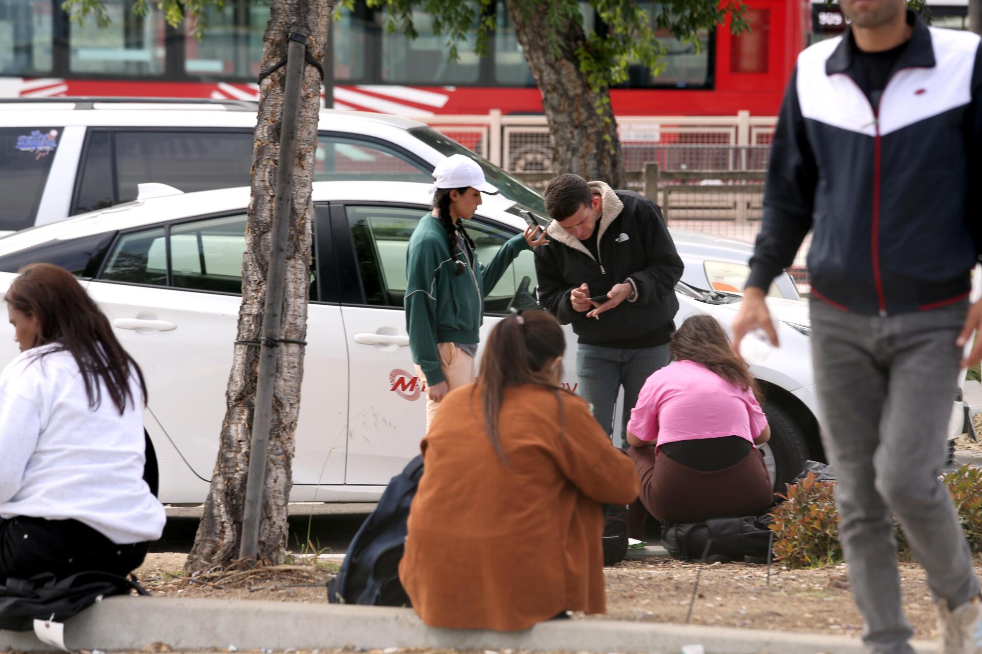 People sit on a sidewalk, some talking to a man.