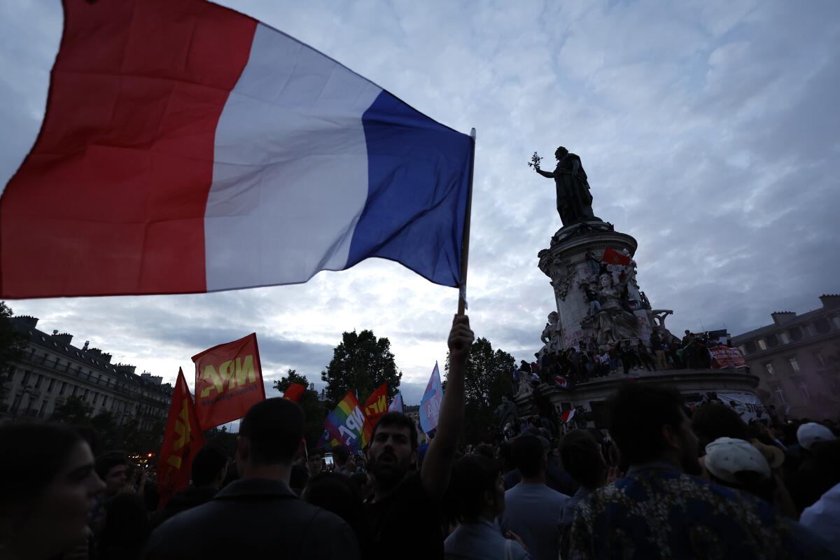 People gather in a plaza, waving a large red, white and blue flag near a monument