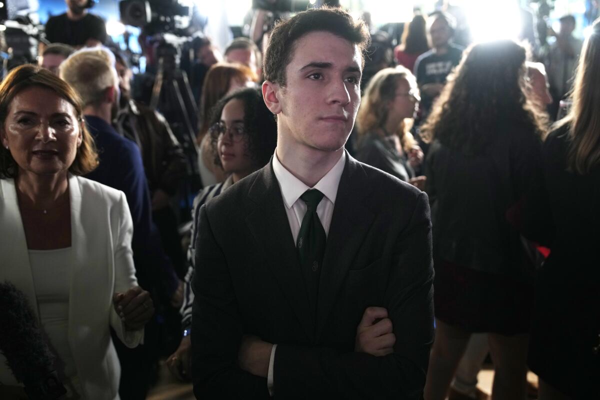 A man in a dark suit and tie, arms crossed, stands next to a woman in a white outfit in the midst of a crowd  