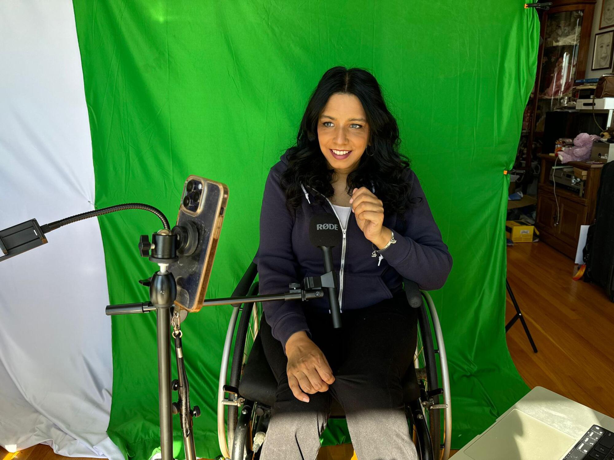 A woman in dark clothes, seated in a wheelchair against a green background, records herself on her cellphone