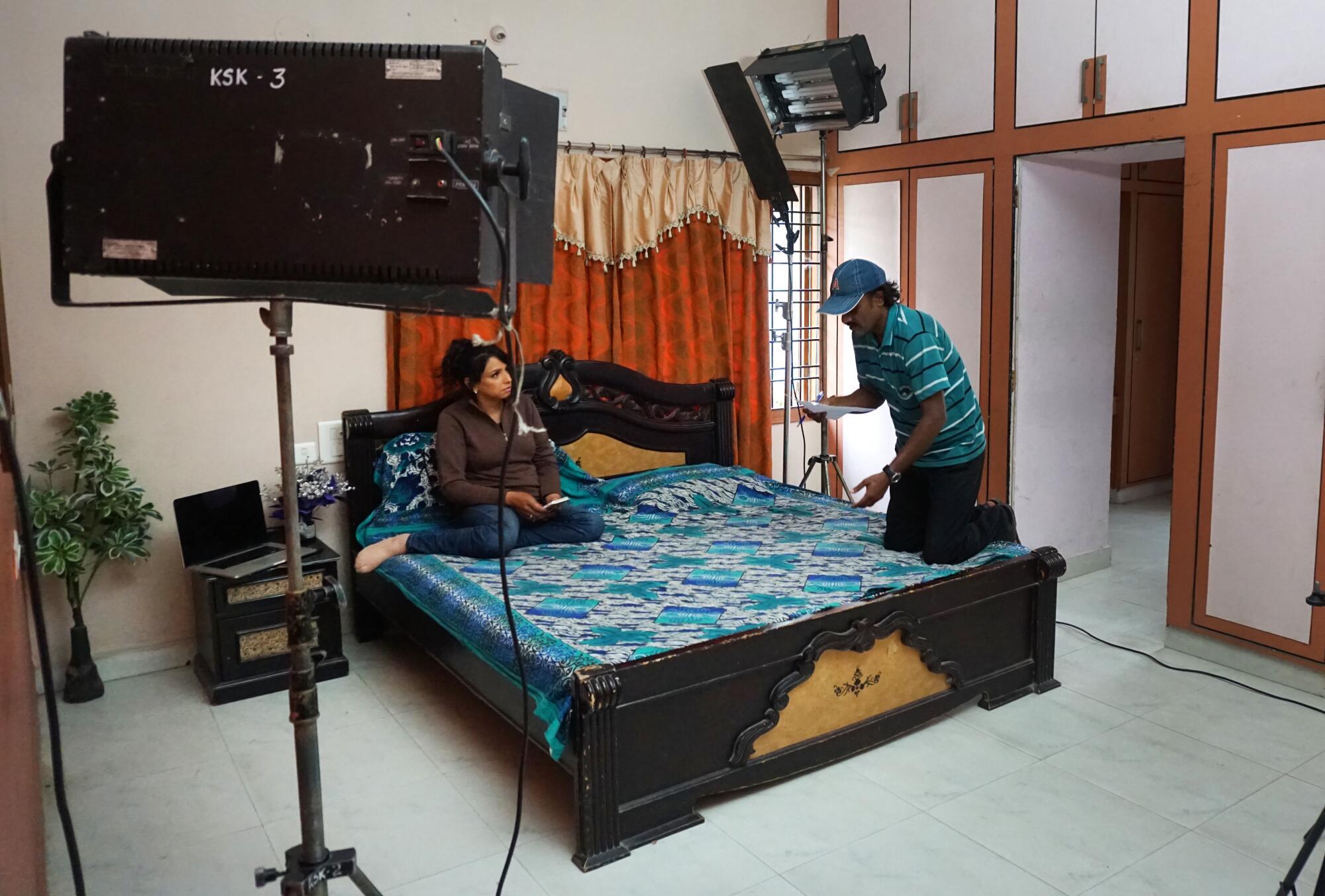 A woman sits on a bed, as a man stands nearby, with recording equipment in the room 