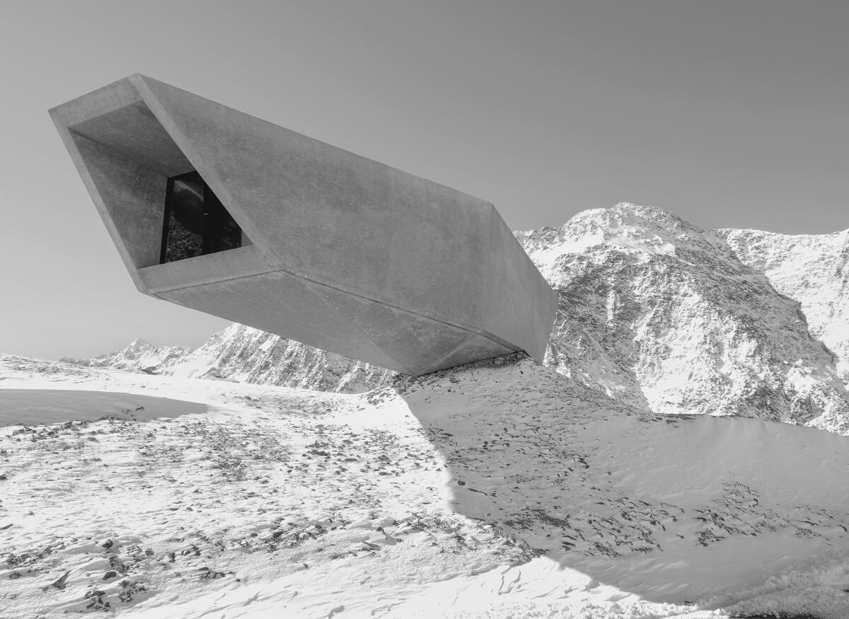 A long, rectangular concrete structure cantilevers out over thin air against the backdrop of snow-covered mountains.