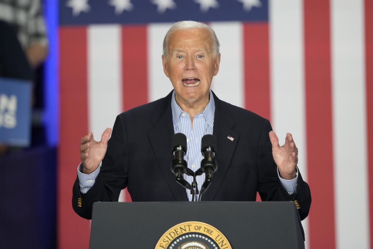 President Biden raising both hands while speaking from a lectern, a large American flag hanging vertically behind him