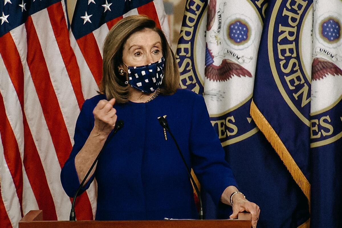 Nancy Pelosi, wearing a fabric mask, raises a clenched hand while speaking, multiple U.S. House and American flags behind her