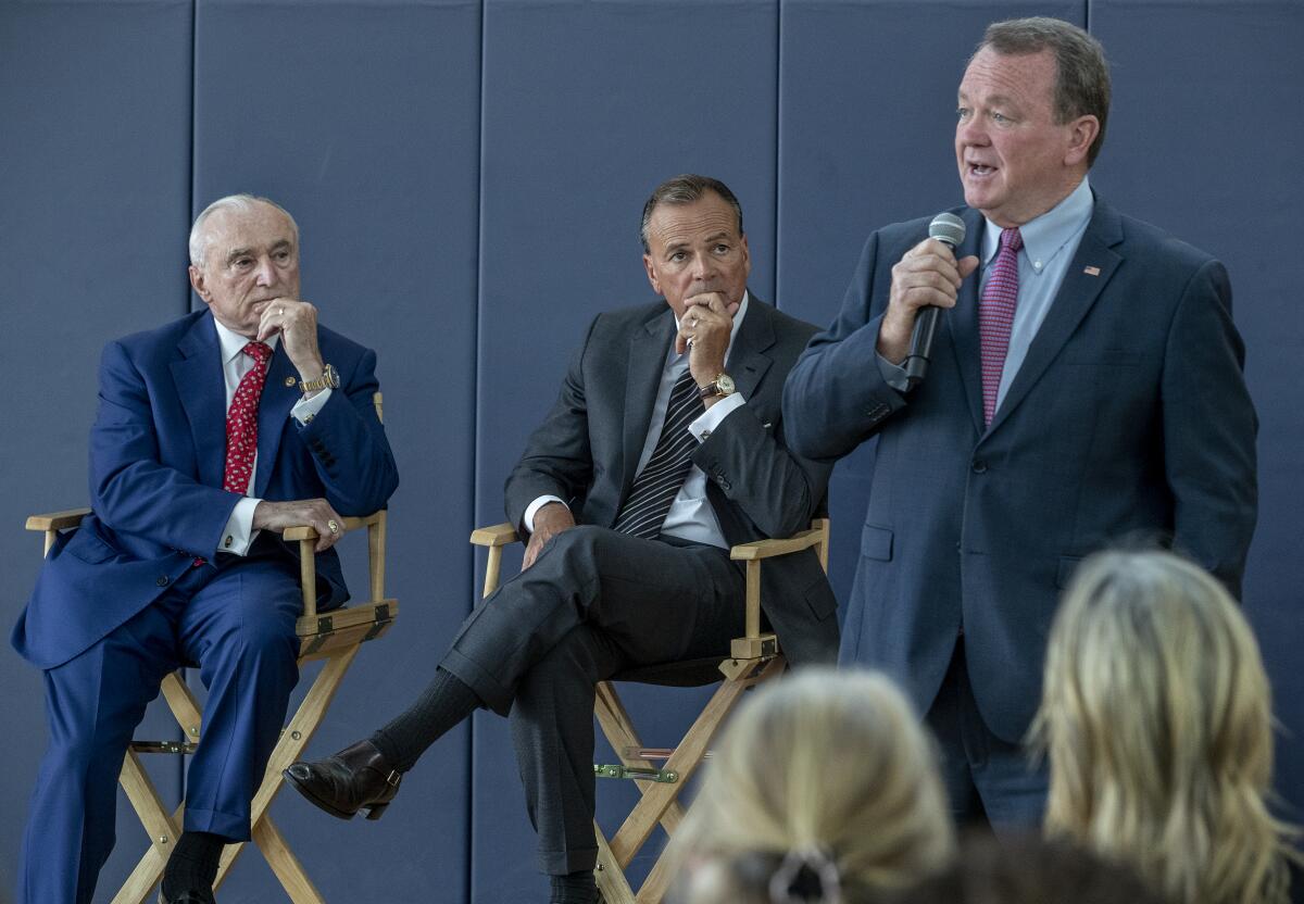 A man stands with a microphone in one hand as two others sit in directors chairs.