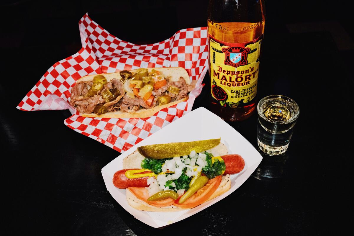 An Italian beef sandwich with a Chicago dog and a bottle and shot of Jeppson's Malört on a black table.