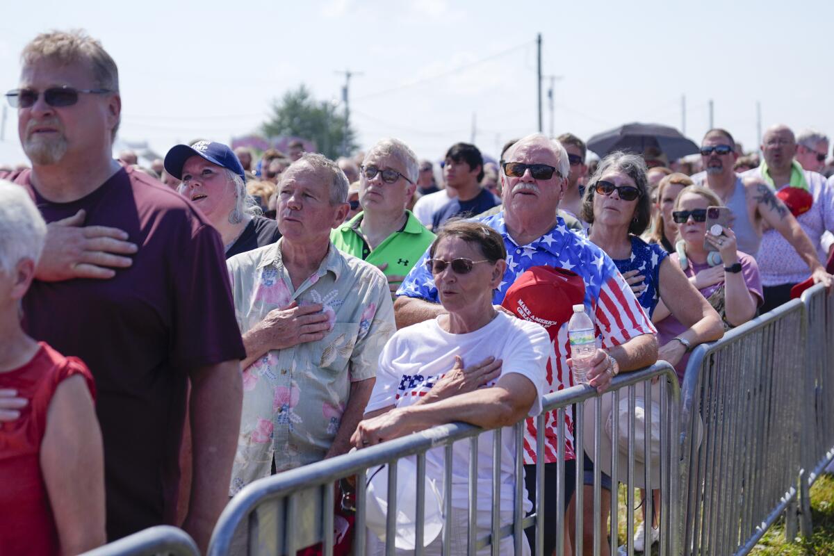 Rally attendees stand near a metal barrier with their right hands on their chests during the Pledge of Allegiance