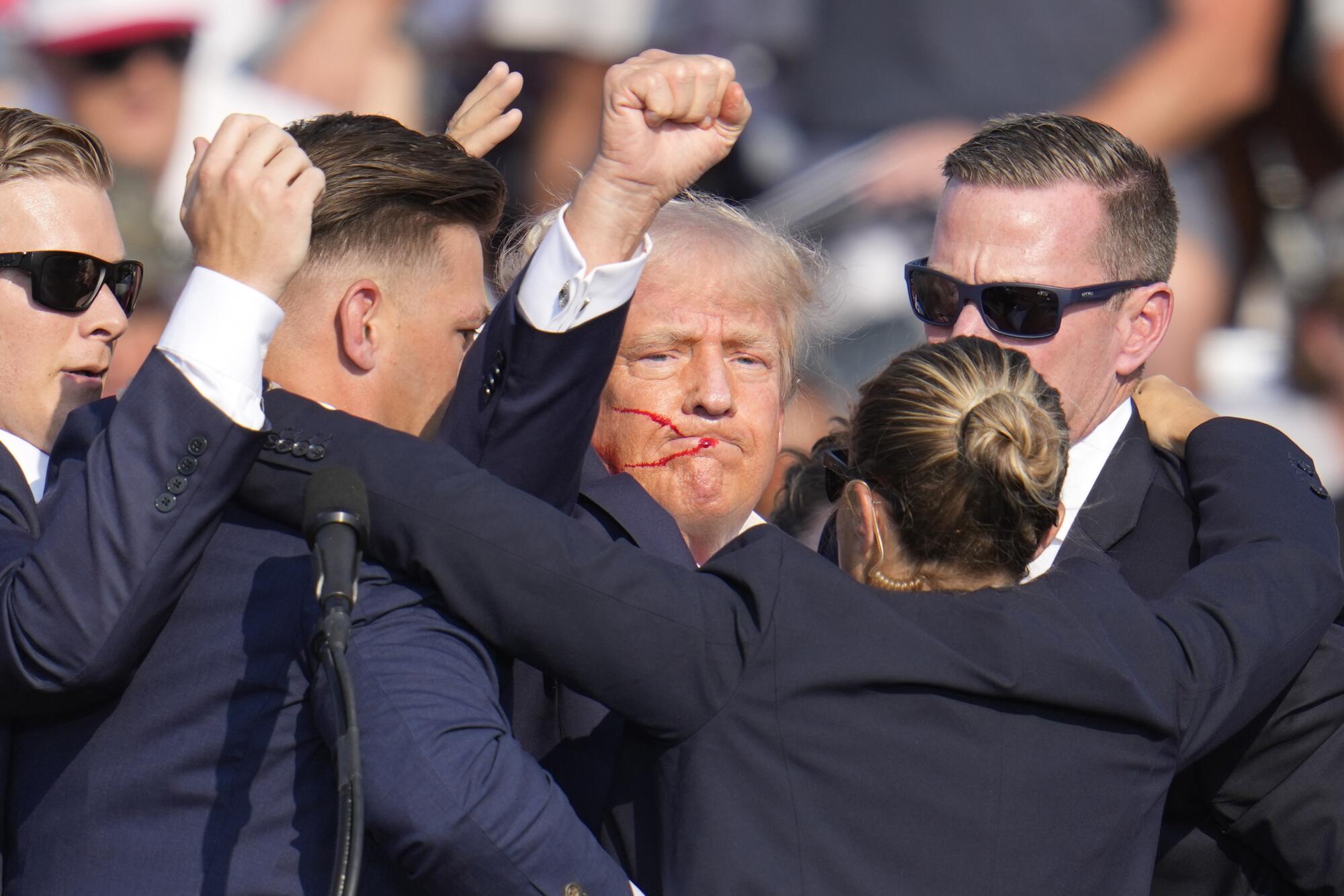 A man surrounded by people in blue suits raises his right fist.