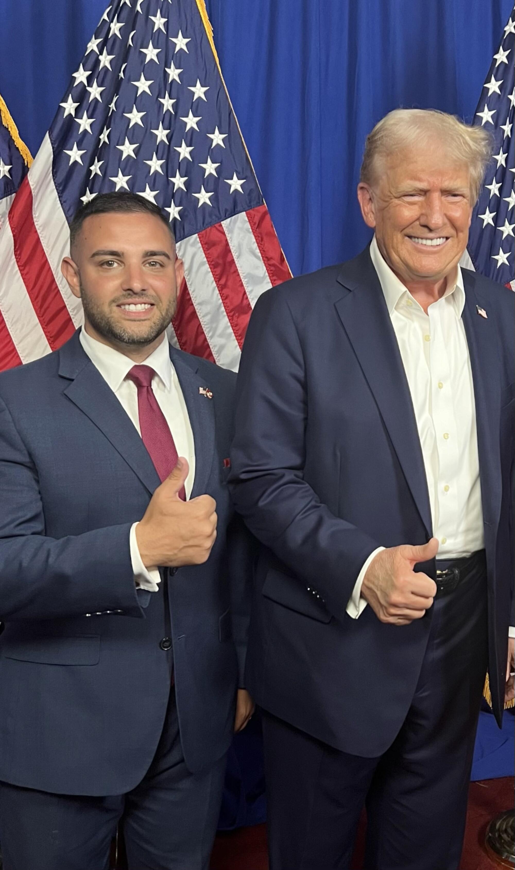 Two men stand in front of American flags.