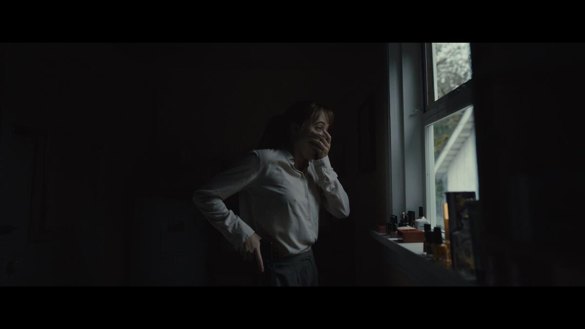 A woman grabs a gun in a holster while looking out a window and covering her mouth with her other hand.