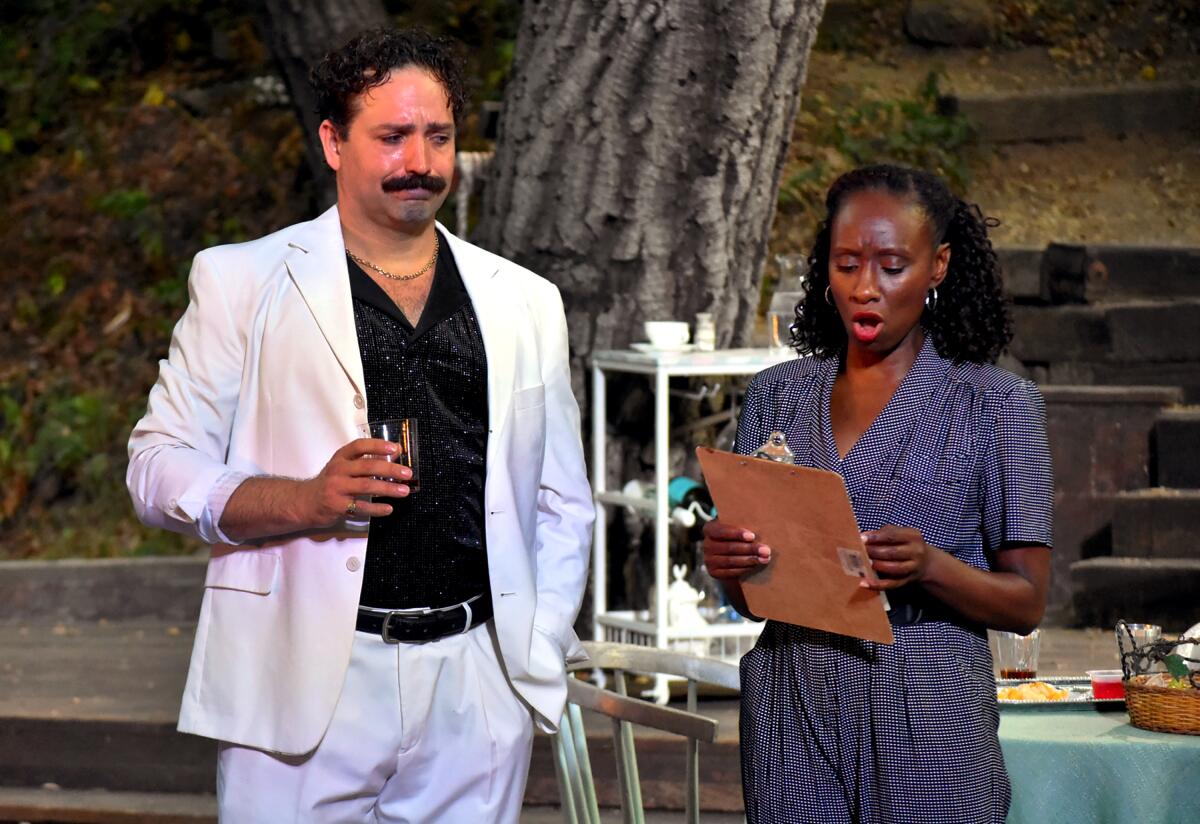 A man in a white suit over dark shirt, holding a drink, stands next to a woman looking at the clipboard she holds